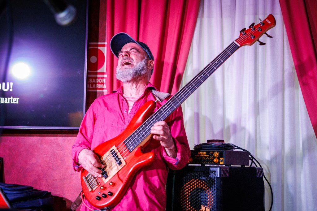 A joyful musician playing a bright red bass guitar, wearing a pink shirt and cap, with an expression of happiness while performing on stage.