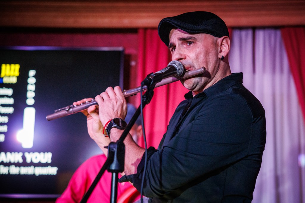 A musician playing the flute on stage, wearing a black shirt and a cap, with a microphone near his mouth and a colorful backdrop behind him.