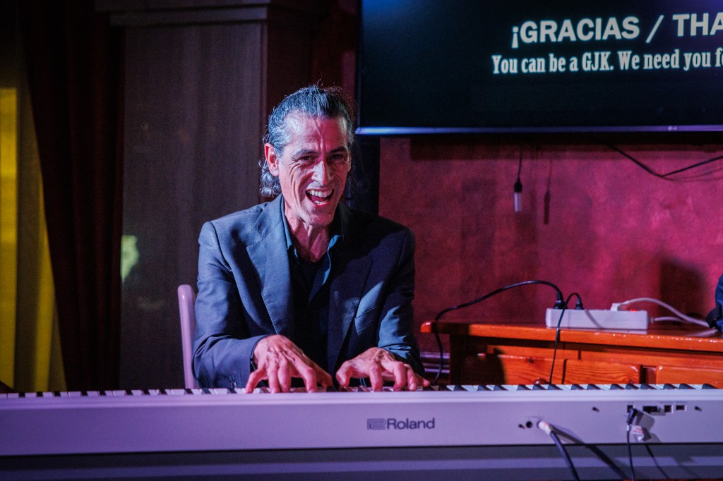 A musician playing a digital piano on stage, smiling and engaged in the performance. The background features warm lighting and a screen with text.