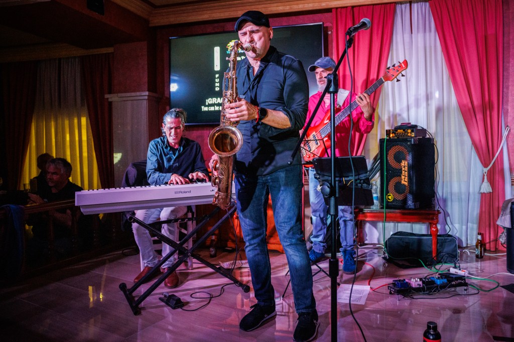 A live band performance featuring a saxophonist in a black shirt, a keyboard player, and a bassist in a red shirt. The setting has a cozy ambiance with curtains and a stage backdrop.