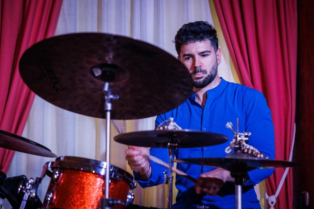 A male drummer playing a drum set, focused and wearing a blue shirt, with curtains in the background.