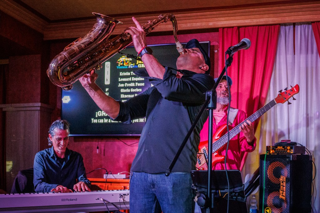A musician playing the saxophone passionately on stage, flanked by a pianist and a bassist. The scene is lively with colorful stage lights and a vibrant atmosphere.