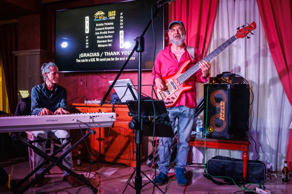 A musician playing a red bass guitar on stage, accompanied by a pianist at a jazz club, with a backdrop displaying 'Gracias / Thank You' and a list of performers.