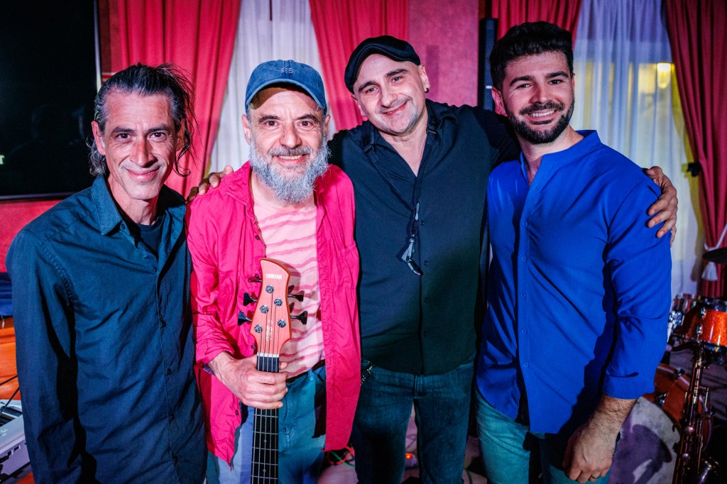 Four musicians posing together, smiling. One holds a guitar, while the others wear casual shirts. The background features musical instruments and a colorful setting.